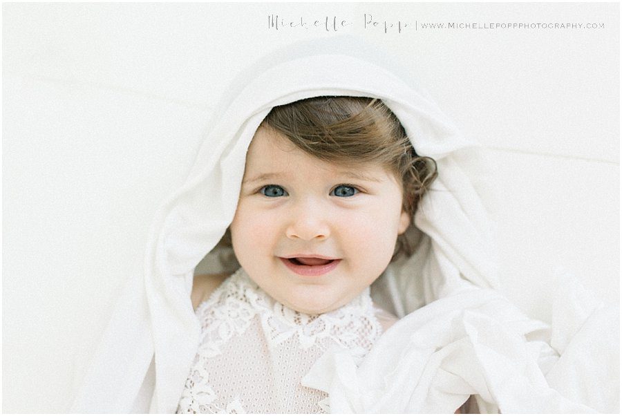 a baby wearing sheets as a veil and smiling while looking at the camera