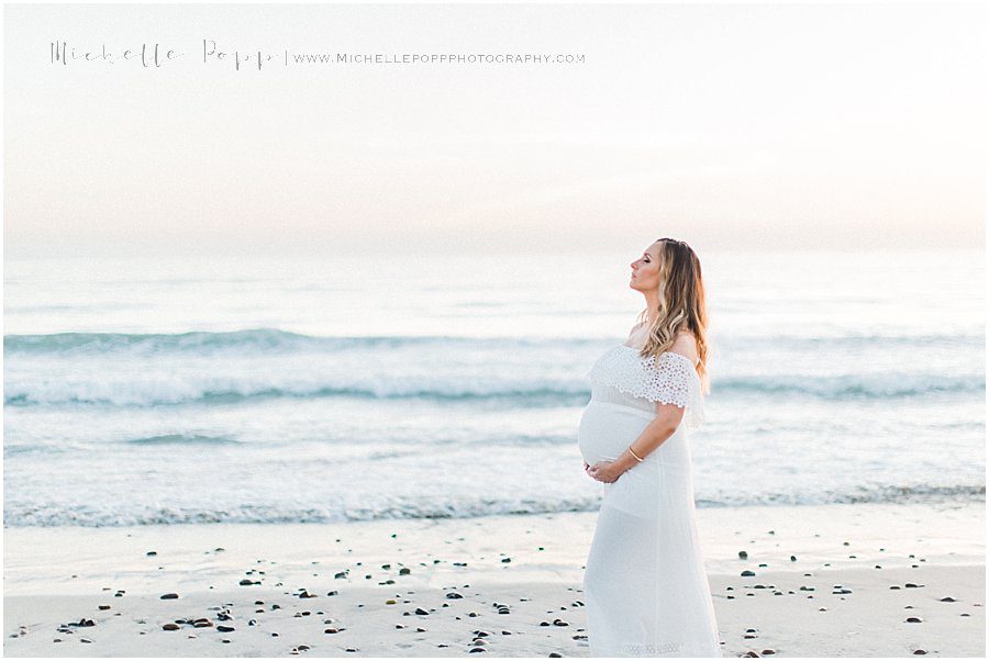 a woman in a white dress looks over the ocean during her maternity photography session