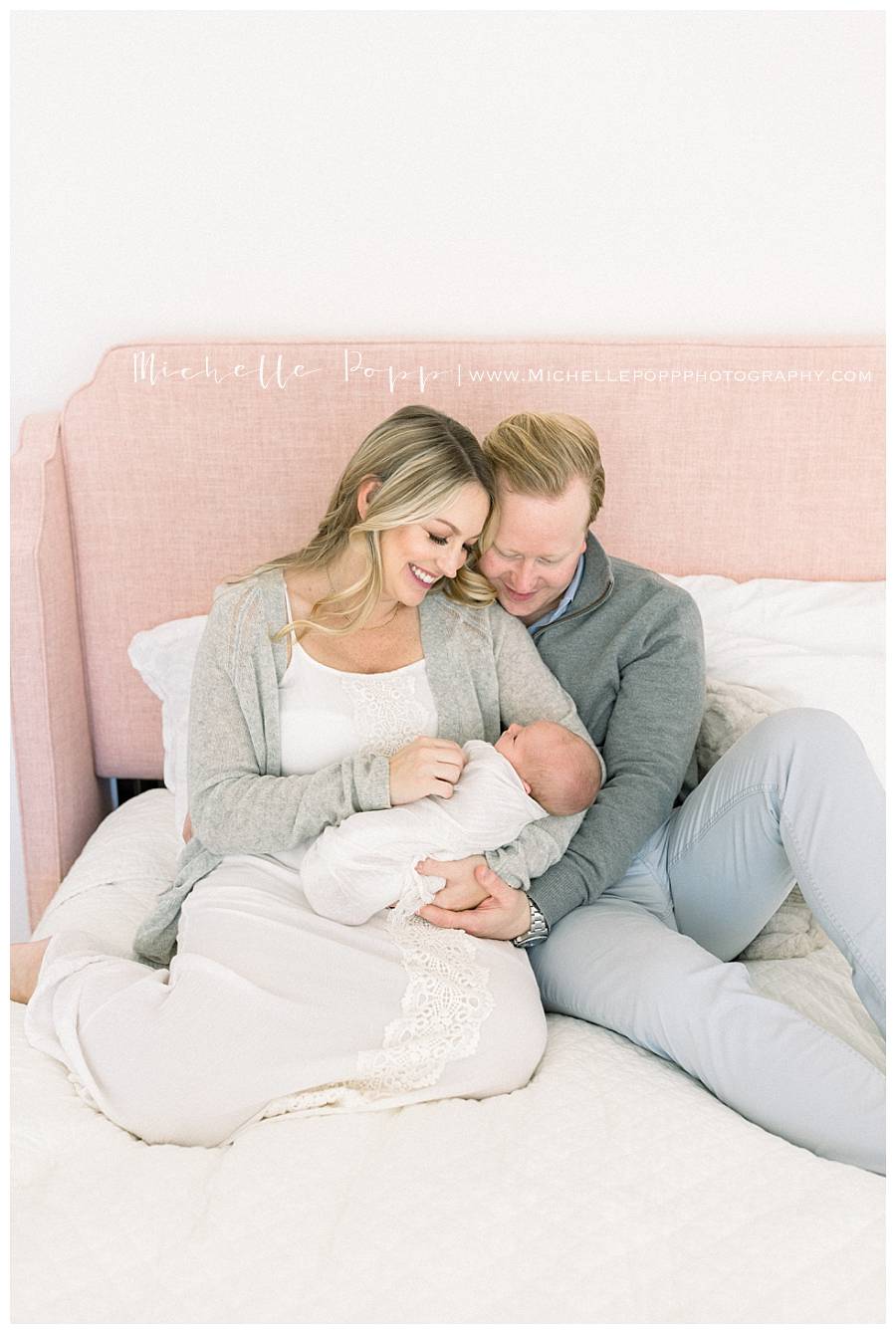 mom and dad sitting on bed looking down at newborn baby