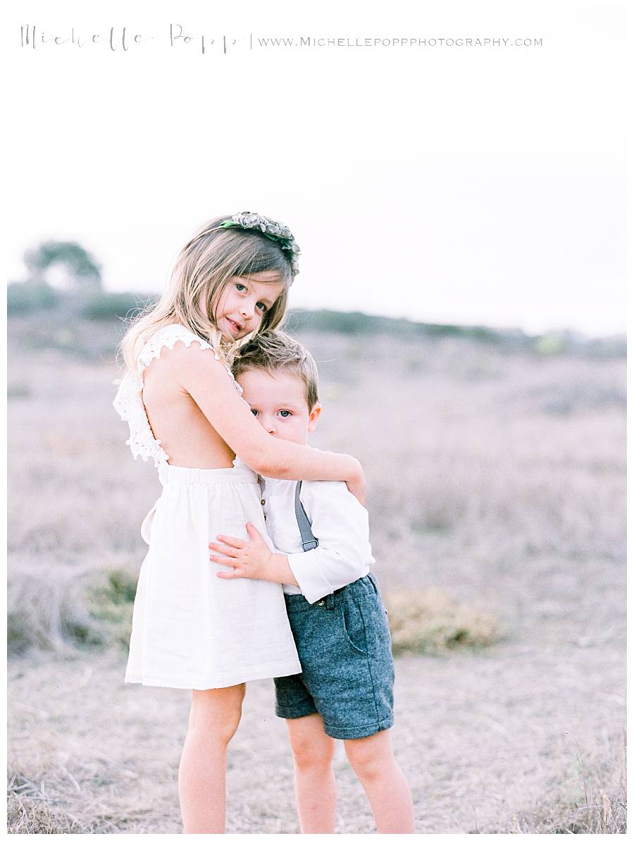little girl in white dress hugging little boy in blue trousers