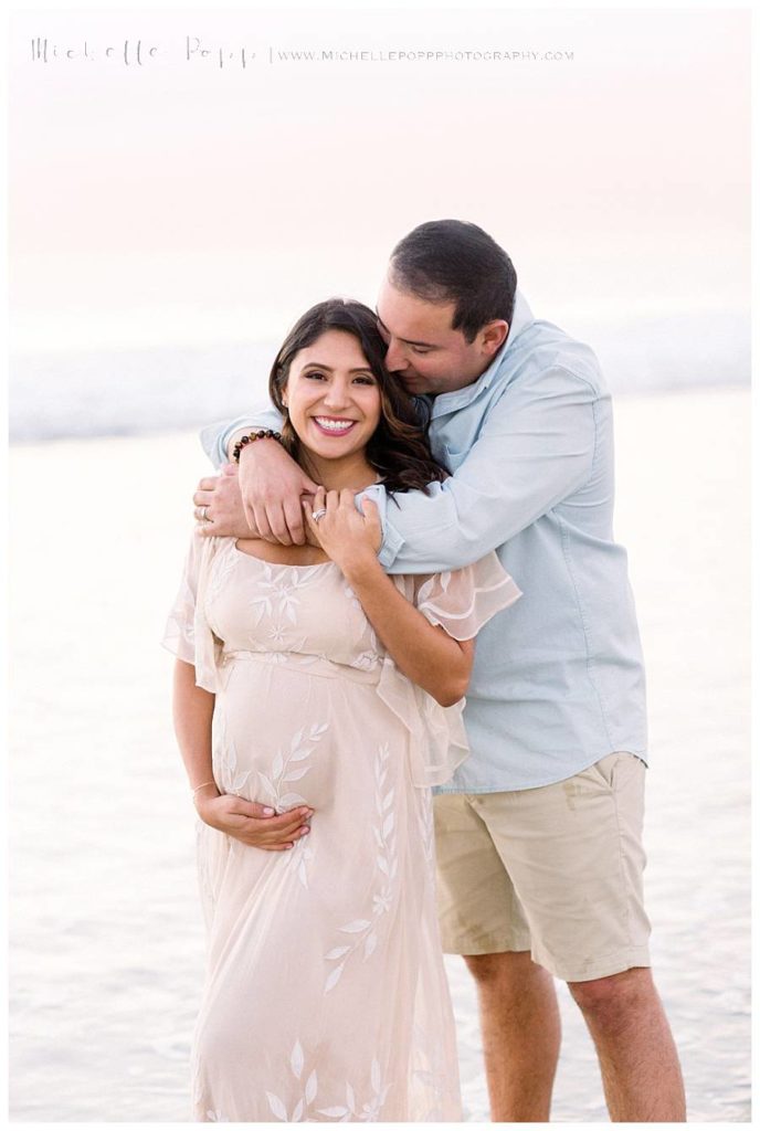 pregnant mom smiling at camera as dad hugs her