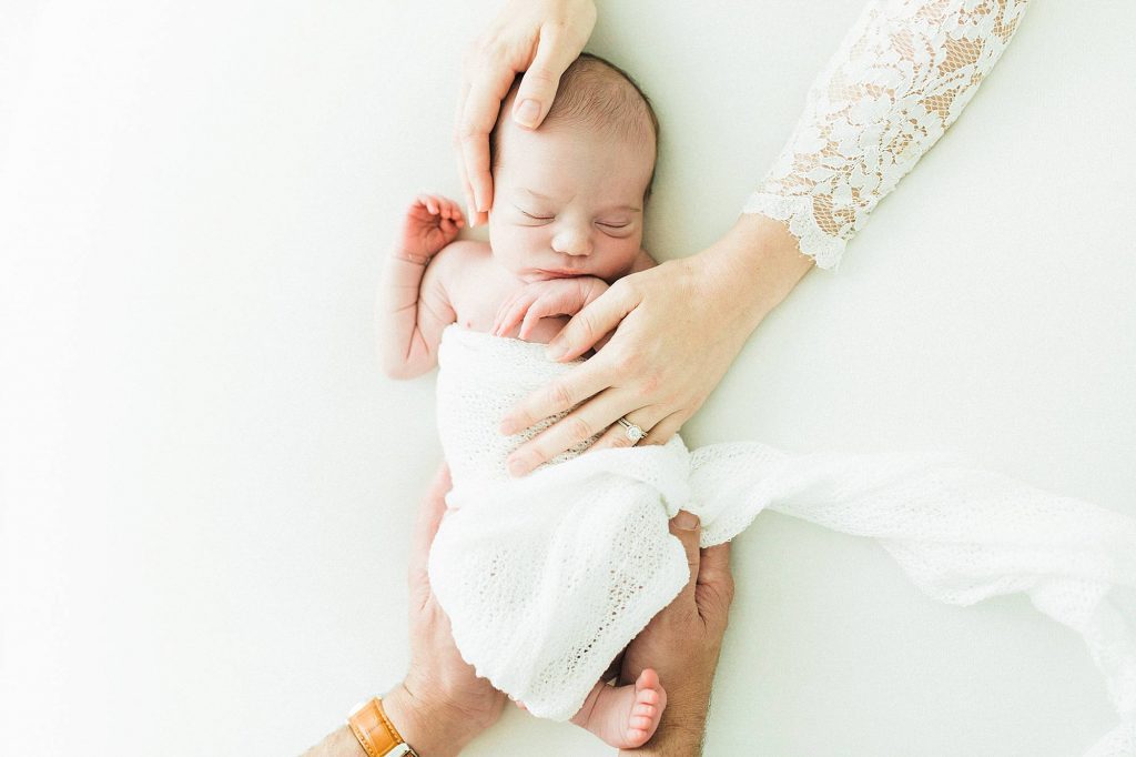 mom and dad's hands on baby sleeping in white swaddle