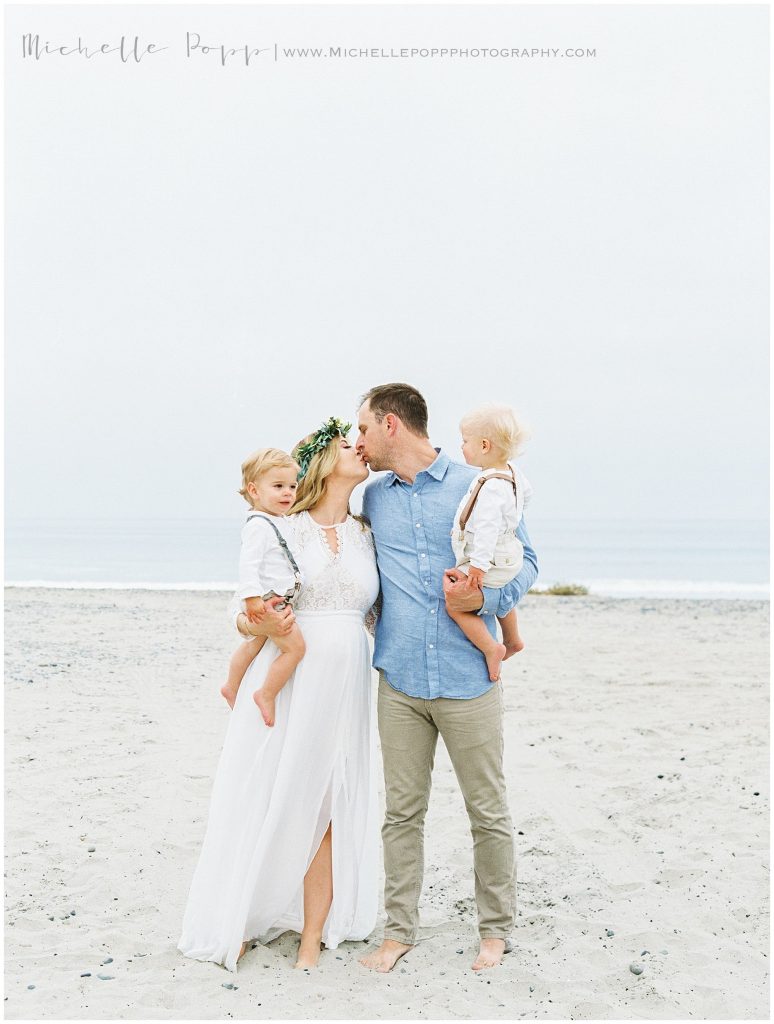 mom and dad kissing on beach while holding boys